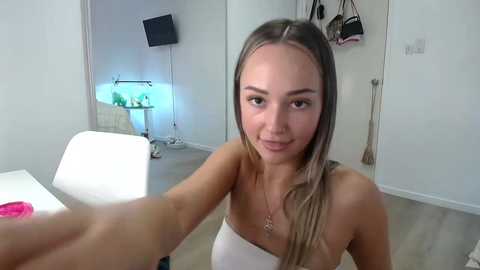 Media: Video of a young, light-skinned woman with long blonde hair, wearing a strapless white top, extending her arm towards the camera in a modern, minimalist room with white walls, a TV, and a white chair.