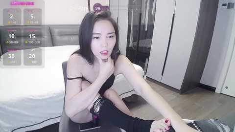 Media: A video of an Asian woman with shoulder-length black hair, wearing a black strapless top and black pants, sitting on a chair in a modern bedroom.