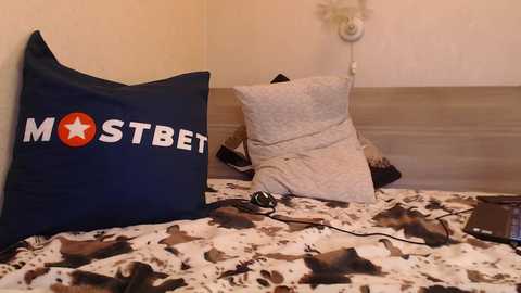 Media: Video of a neatly made bed with a blue pillowcase featuring the word \"MOSTBET\" in white, a white pillow, and a patterned bedspread with brown and white hues.