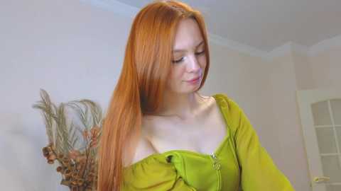 Media: Video of a fair-skinned, red-haired young woman in a lime-green off-shoulder top, standing indoors near a vase of dried flowers and a white door.