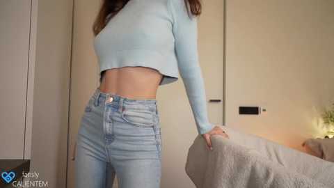 Media: A video of a young woman with light skin, wearing a light blue, long-sleeve crop top and high-waisted light blue jeans, leaning against a beige door in a minimalist bedroom.