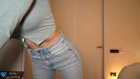 Media: Video of a person wearing a grey crop top and light blue jeans, standing indoors with beige walls and a closed door in the background.