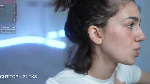 Media: Video of a young woman with light skin and dark hair in a ponytail, wearing a white t-shirt. She appears to be in a room with a blurred blue and white backdrop. Text at the bottom reads \"CUTTOR-21-TKS.\