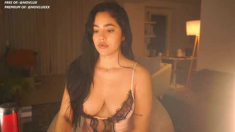 Media: A video of a young, curvy woman with long black hair, wearing a revealing pink lace lingerie top, standing in a dimly-lit living room with a lamp and a glass of red drink.