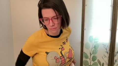 Media: Video of a woman with short, dark hair, wearing glasses and a yellow T-shirt with a cartoon character, standing in a room with a floral-patterned shower curtain.