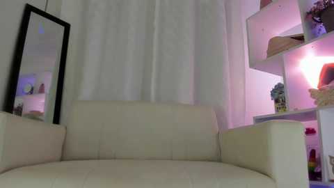 Media: A video of a minimalist, modern living room with a cream sofa, white curtains, and a black-framed mirror. A white bookshelf with pink lighting and decorative items is visible in the background.