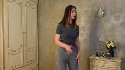 Media: Video of a young woman with long brown hair, wearing a gray T-shirt and jeans, standing in a beige, ornate room with a wall-mounted lamp, white wardrobe, and a small table with a vase of flowers.