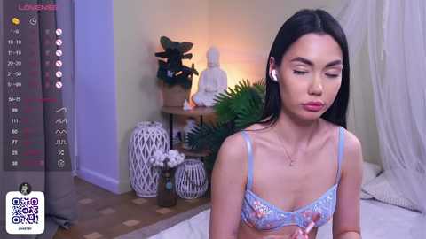 Media: Video of an Asian woman with long black hair, wearing light blue floral lingerie, sitting on a bed with white pillows, in a cozy bedroom with a Buddha statue, plants, and a digital timer screen.