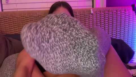 Media: Video of a woman with shoulder-length brown hair, wearing a gray sweater, lying on a wicker couch with a purple light illuminating the background.