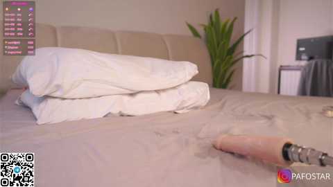 Media: Video of a woman's hand holding a realistic sex toy, on a neatly made bed with white sheets and a beige headboard. The background features a potted plant and a TV, creating a modern, minimalist bedroom setting.