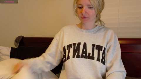 Media: A video of a blonde woman with fair skin wearing a gray sweatshirt with \"MICHIGAN\" in bold letters, sitting on a bed with beige walls and a wooden headboard.