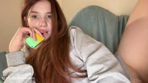 Media: A video of a young, fair-skinned woman with long, straight, auburn hair, wearing a light gray hoodie, lying on a blue couch, holding a green and yellow smartphone to her mouth, with a blurry, partially visible person in the background.