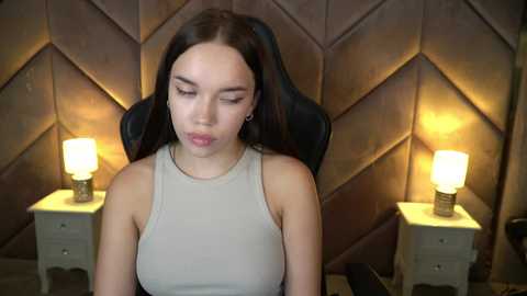 Media: Video of a young woman with long brown hair, wearing a light gray sleeveless top, sitting in a black gaming chair with diamond-patterned, brown upholstered walls in the background, featuring two beige nightstands with lit lamps.