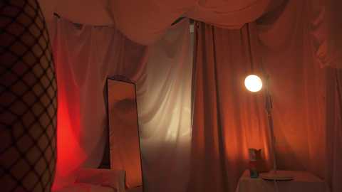 Media: Video of a dimly lit, intimate bedroom with sheer curtains, a standing mirror, a round lamp, and a bed with a quilted cover. Red and orange hues dominate the room, creating a warm, cozy atmosphere.