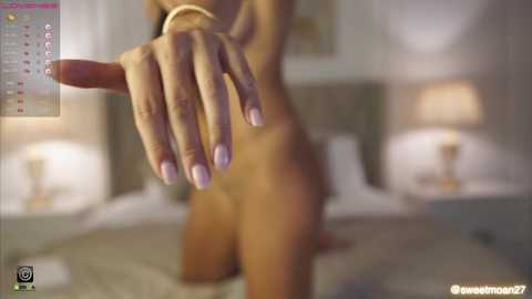 Media: A blurry, dimly lit video of a nude person with a tan complexion, wearing a gold bracelet, standing near a bed with white sheets and two lamps. The image has a watermark \"sweetmom22\" in the bottom right corner.