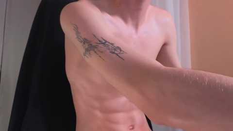 Media: Video of a shirtless, fair-skinned man with a defined, muscular physique, showcasing a black, abstract tattoo on his right upper arm. Background features a black towel and a beige wall, creating a casual indoor setting.