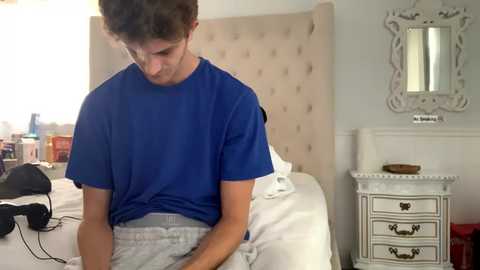 Media: Video of a young man with short curly hair in a blue T-shirt and gray sweatpants sitting on a bed, adjusting his clothes in a cozy, light-colored bedroom.