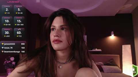 Media: Video of a young woman with long brown hair and fair skin, wearing a black necklace, sitting in a dimly lit bedroom with a white lamp and a bed in the background.