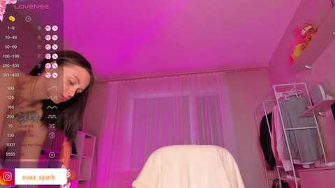 Media: Video of a woman with long black hair and light skin, wearing a beige top, leaning forward in a pink-lit room.