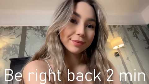 Media: Video of a young woman with wavy, blonde hair and light skin, smiling, wearing a white tank top, in a room with a beige, tree-patterned wallpaper and a modern lamp. Text reads, \"Be right back 2 min.\