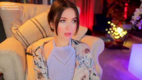 Media: Video of a young woman with fair skin and brown hair, wearing a floral-patterned cardigan over a light blue sweater, seated in a beige armchair in a dimly lit living room with warm lighting and a festive atmosphere.