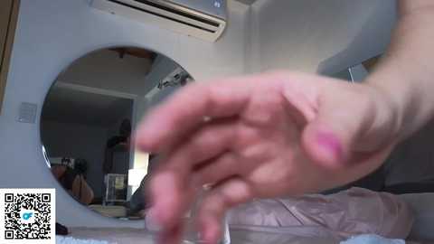 Media: Video of a person's hand with pink nails reaching into a washing machine. The background shows a partially visible bedroom with a bed, air conditioner, and mirror.