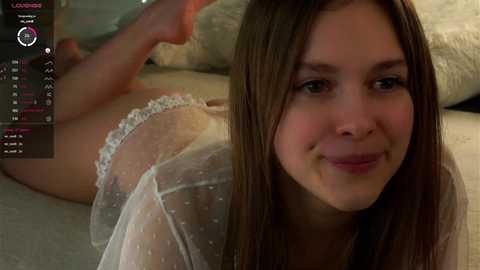 Media: Video of a young woman with fair skin and straight brown hair, lying on her stomach in white polka-dotted lingerie. She has a soft smile. Background includes a beige couch and pillows.