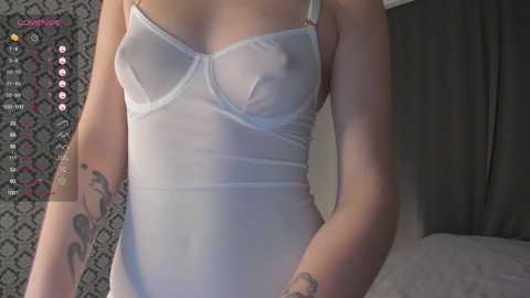 Media: Video of a slender, light-skinned woman wearing a sheer white lingerie bodysuit, revealing her nipples and tattoos on her arms. Background features a dark curtain and patterned wallpaper.