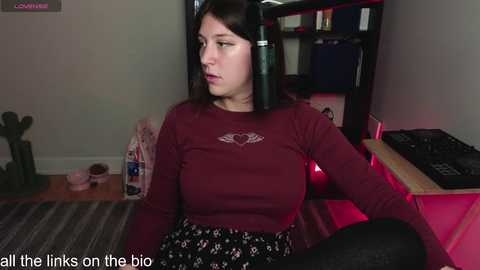 Media: Video of a young woman with long dark hair, wearing a maroon long-sleeve shirt and black floral skirt, sitting on the floor in a dimly lit room with a bookshelf and gaming equipment.