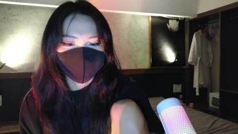 Media: A video of a young Asian woman with long black hair, wearing a black face mask, a black top, and holding a white and blue disposable cup, in a dimly lit bedroom with wooden furniture and a white robe hanging on a door.