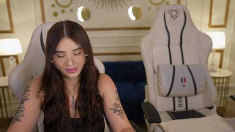 Media: Video of a woman with long, wavy brown hair, wearing a black tank top, seated in a white gaming chair with a grey accent. She has multiple tattoos and is focused on her phone, with a modern, warmly lit room in the background.