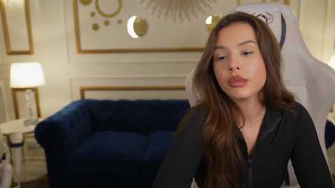 Media: Video of a young woman with long brown hair, wearing a black top, seated in a stylish, dimly-lit room with a blue velvet sofa and modern art on the wall.