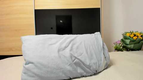 Media: Video of a light blue pillow on a white bed, with a wooden wardrobe and a bouquet of yellow flowers in the background.