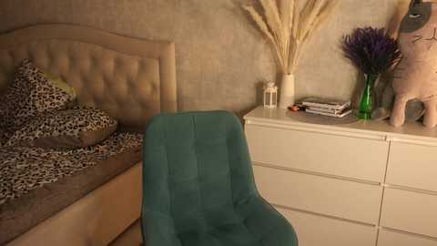 Media: A video of a modern bedroom featuring a teal chair, a bed with leopard print bedding, a beige tufted headboard, a white dresser with a green vase, and a beige wall.