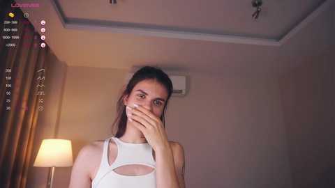 Media: Video of a young woman with long brown hair, wearing a white tank top, covering her mouth, standing in a dimly lit room with beige walls and a white ceiling fan.