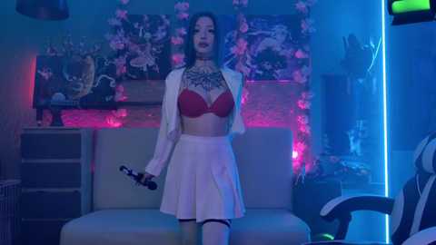 Media: Video of a tattooed woman in a red bra and white skirt, holding a controller, in a dimly lit, pink-lit room with gaming furniture.