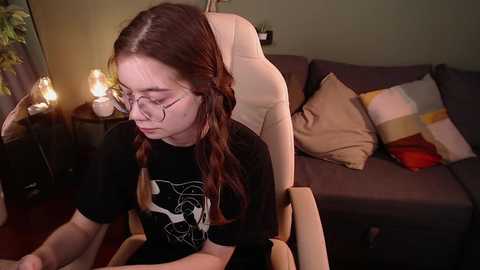 Media: Video of a young woman with glasses and long brown hair in braids, wearing a black T-shirt, seated in a beige chair, surrounded by warm lighting and patterned cushions in a cozy living room.