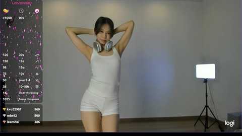 Media: Video of an Asian woman with fair skin, dark hair, and a slim build, wearing white shorts and a tank top, standing with hands behind head, surrounded by streaming platform interface.