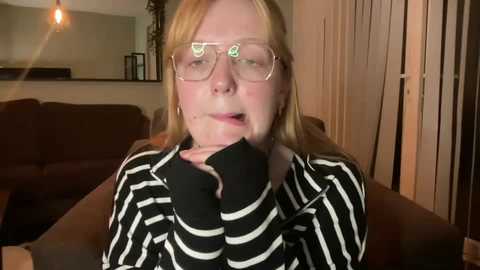 Media: Video of a woman with fair skin, blonde hair, and glasses, wearing a black and white striped shirt and black gloves, sitting on a brown couch, in a dimly lit living room with vertical blinds and a brown coffee table.