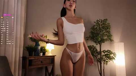 Media: Video of an Asian woman with long black hair, wearing a white crop top and sheer white panties, standing in a softly lit, beige-walled bedroom.