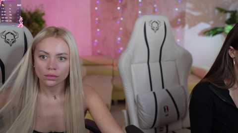 Media: Video of a young blonde woman with fair skin, wearing a black top, sitting on a white gaming chair in a cozy room with pink fairy lights and a green plant.