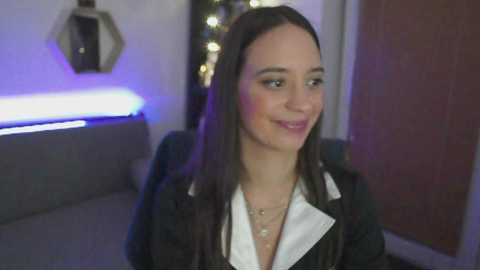 Media: A video of a young woman with long brown hair, wearing a black jacket over a white blouse, smiling in a dimly lit room with a hexagonal mirror and glowing blue lights in the background.