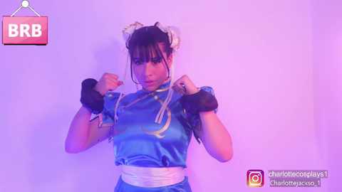 Media: Video of a woman in a Chun-Li cosplay, featuring a blue qipao dress, white gloves, and a white headband. She poses confidently with a purple background, with the \"BRB\" logo in the top left corner.