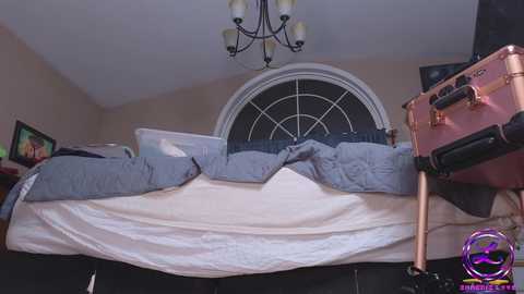 Media: Video of a cozy bedroom featuring a bed with white and blue bedding, a circular window, a chandelier, and a purple logo watermark.