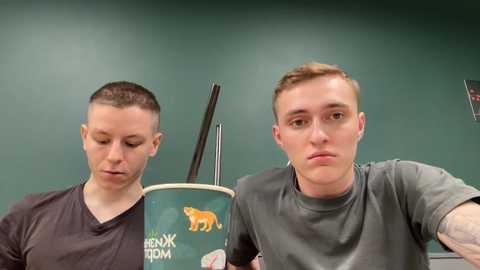 Media: Video of two young men with short hair, one wearing a gray shirt, the other a black shirt, seated at a table with chopsticks and a green cup featuring a cartoon tiger, in a green-walled room.