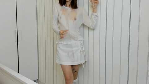 Media: Video of a slender woman with fair skin, shoulder-length dark hair, wearing a sheer white top and mini skirt, posing against vertical white blinds in a minimalistic room.