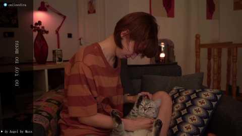 Media: Video of a young woman with short brown hair, wearing a striped shirt, cuddling a kitten on a bed with a blue patterned blanket. Room has wooden furniture, red lamp, and framed pictures.