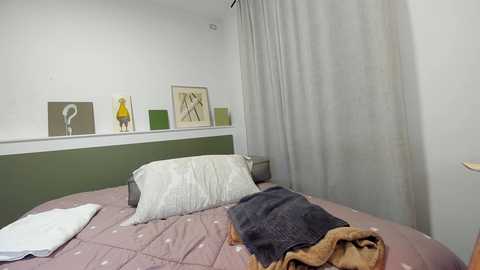 Media: A video of a minimalist bedroom with a green headboard, beige bedspread, white pillow, and a beige blanket. The wall features three abstract artworks in green, yellow, and white. A sheer white curtain partially covers the window.