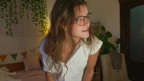 Media: Video of a young woman with curly brown hair, wearing glasses, a white T-shirt, and smiling, standing in a cozy bedroom with green ivy wall decor, a potted plant, and a bed with geometric-patterned pillows.