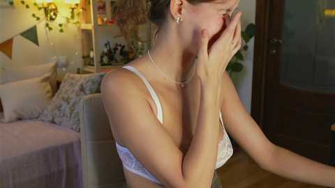 Media: Video of a light-skinned woman with brown hair in a white lace bra, covering her face with her hands, sitting in a dimly lit bedroom with a bed, wall decorations, and a door.